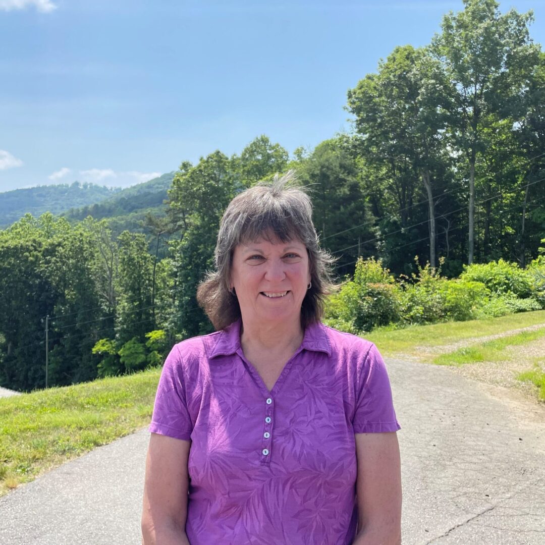 Person in a purple shirt standing on a path with trees and hills in the background on a sunny day.