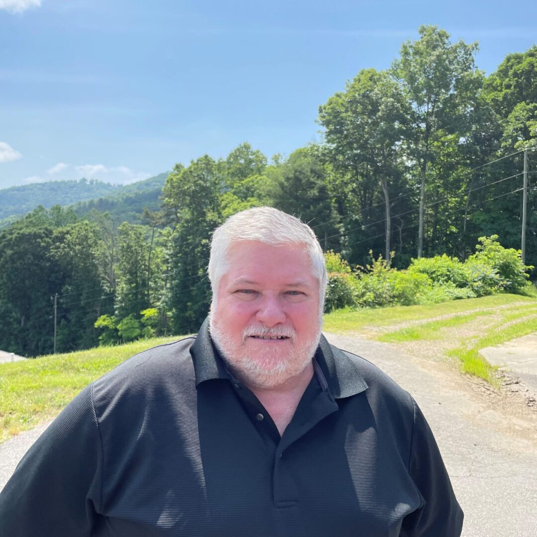 A person with short gray hair and a beard stands outdoors in front of a hilly, tree-filled background on a sunny day.