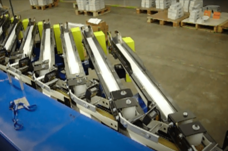 Conveyor belts with boxes in a warehouse setting, with pallets in the background.